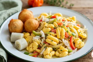 Ackee and saltfish on a white enamel plate with a dark green rim: soft yellow ackee lobes with flakes of salt fish, onion, scallion, red and green pepper, tomato and thyme, three fried dumplings and two pieces of boiled green banana, a whole Scotch bonnet behind