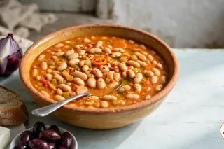 A deep ochre-glazed earthenware bowl of Greek fasolada on a pale blue wooden table: plump white beans in a thick orange-red broth with carrot rounds, celery, a sheen of olive oil and chili flakes, a spoon in the bowl, with country bread, feta with oregano, Kalamata olives and a quartered red onion beside it