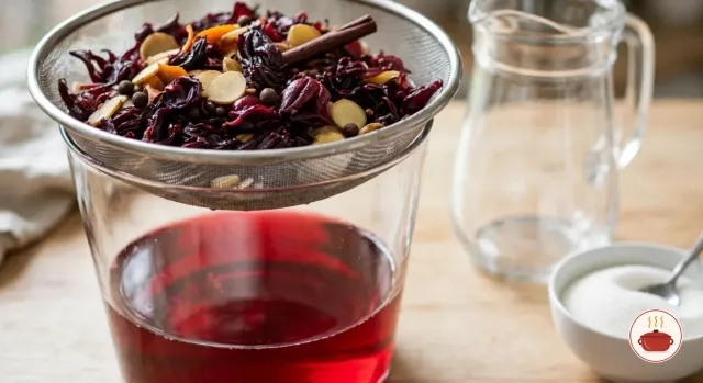 Spent, dark burgundy sorrel calyces with ginger coins, a cinnamon stick and pimento berries in a fine sieve resting high over a tall glass bowl of strained ruby-red sorrel, with an empty glass pitcher and a small bowl of sugar beside it