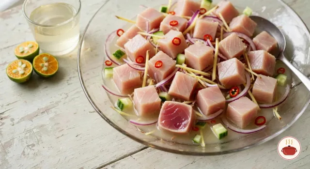 Cubes of tuna turned pale pink outside by the vinegar in a wide glass bowl, one halved cube showing a translucent red center, with red onion, ginger matchsticks, chili rings and diced cucumber in a little cloudy dressing, calamansi halves beside it