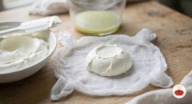 A dense mound of strained labneh on an opened white cheesecloth, more labneh already scraped into a white bowl beside it with a white spatula on the rim, and the drained whey in a glass bowl behind