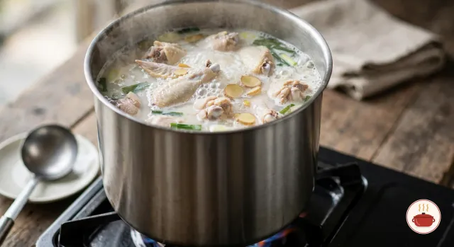 Bone-in chicken pieces in a tall stainless-steel stockpot at a hard, rolling boil on a gas burner, the broth already cloudy and milky white with bubbles breaking across the surface, a few slices of ginger and green onion tops among the chicken