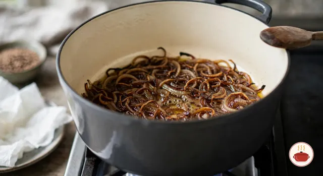 Thinly sliced onions fried to a deep chestnut brown in a thin film of olive oil on the bottom of a charcoal-grey enameled Dutch oven on a gas burner, with a wooden spoon resting on the rim and a plate lined with paper towel beside the stove
