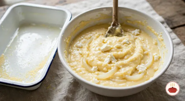 A wide white bowl of thick, creamy, pale yellow cornmeal batter with crumbles of white cheese and soft onion slices folded through it, a wooden spoon standing in it, beside the empty white enamel pan, greased and dusted with cornmeal