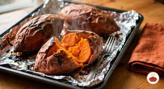 Sweet potatoes just baked in their skins on a foil-lined dark baking sheet, the copper-red skins wrinkled and slumped, dark caramelized syrup leaking onto the foil, the front one split open to show soft, steaming deep-orange flesh, with a fork and a rust-orange napkin beside the pan