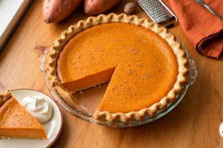 A homemade sweet potato pie in a clear glass pie plate on a honey-colored pine table: a smooth, matte burnt-orange filling flecked with nutmeg inside a fluted golden crust, one wedge cut out and set on a cream plate with whipped cream, with raw copper-skinned sweet potatoes, a whole nutmeg, a grater and a rust-orange napkin behind it