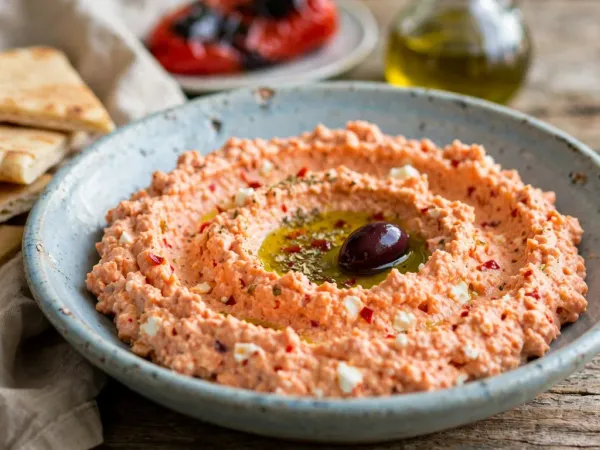 Greek tirokafteri in a shallow, speckled blue-grey stoneware bowl: a thick, coral-orange spread of whipped feta with small white crumbs of feta and flecks of red chili, swirled around a pool of olive oil with dried oregano and a Kalamata olive, beside warm pita triangles, with a roasted red pepper and a cruet of olive oil behind