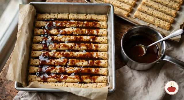 One layer of pale golden turrón sticks laid side by side in a parchment-lined square pan and glazed with dark amber syrup, next to a small steel saucepan of syrup with a spoon and a baking sheet of more sticks
