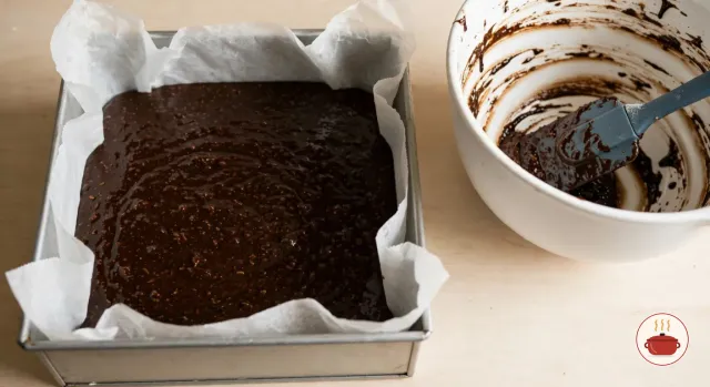Dark, glossy, oat-flecked parkin batter freshly poured into a square cake tin lined with baking parchment, beside a white mixing bowl streaked with batter and a spatula